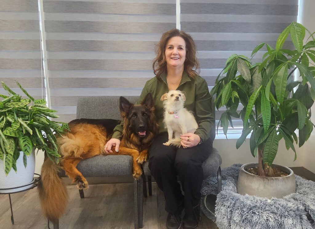 A woman sits on a chair with a German Shepherd lying beside her and a small dog on her lap, surrounded by indoor plants.